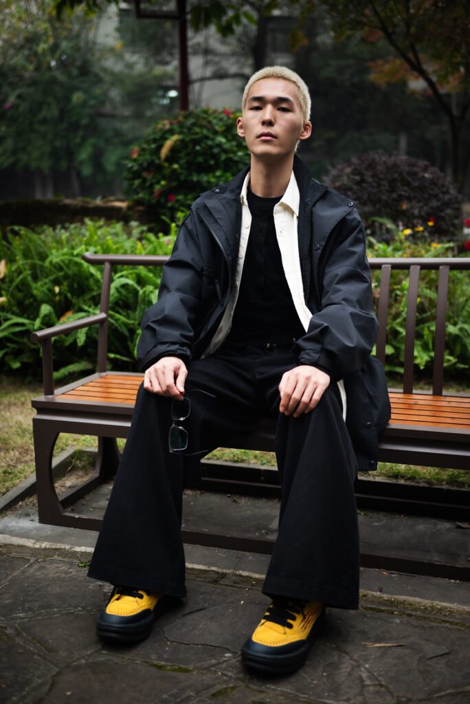 Young Asian man in stylish black outfit sitting on park bench.