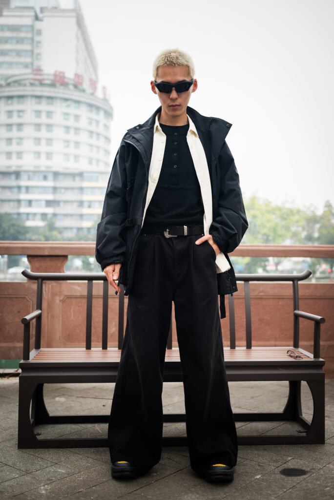 Young man in black suit and sunglasses standing in front of a street bench.