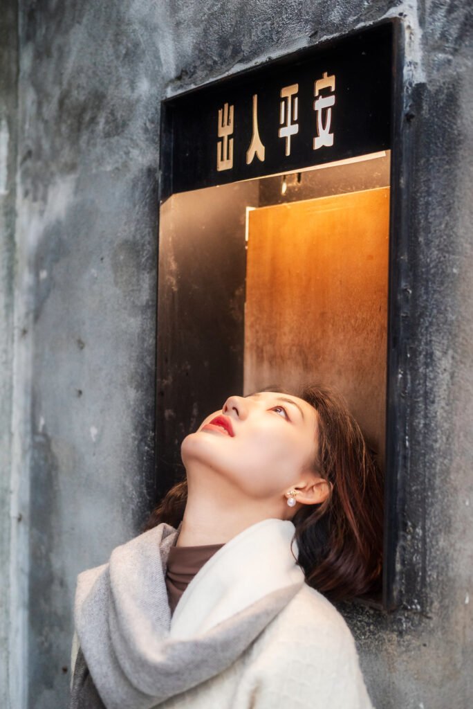Young woman in a white sweater and gray scarf standing in front of a door.