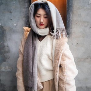 Young woman in a cream-colored coat and gray scarf standing in front of a gray wall.