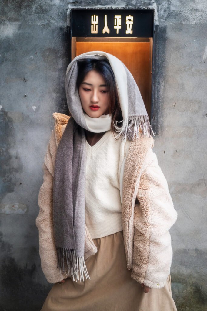 Young woman in a cream-colored coat and gray scarf standing in front of a gray wall.