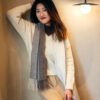 Young woman standing by the wall and leaning against it with one hand while the other is holding the end of a long scarf.
