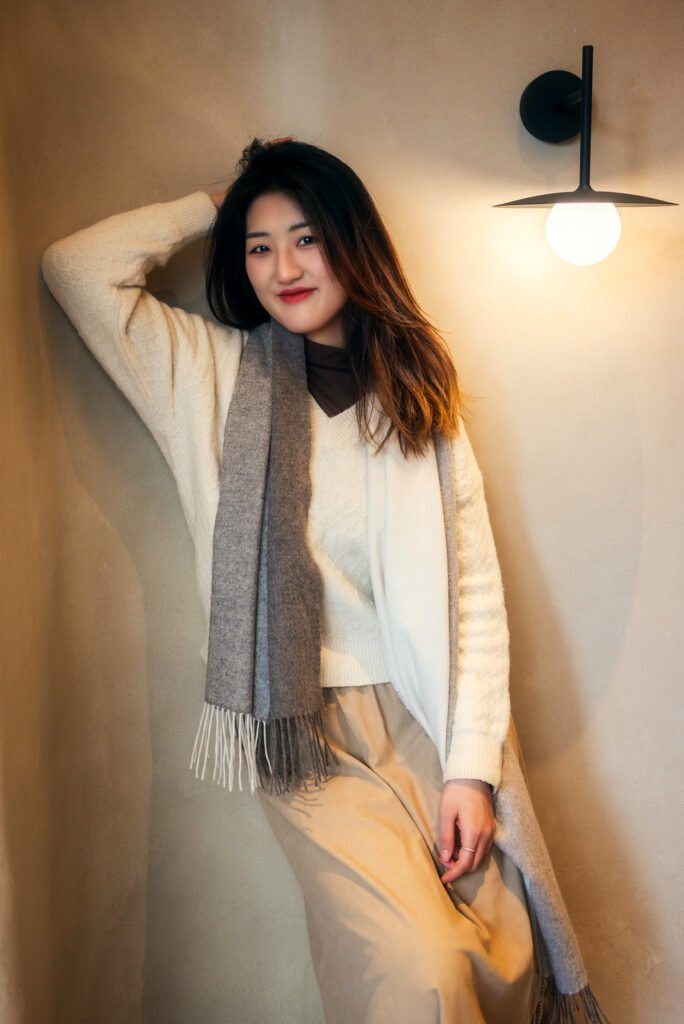 Young woman standing by the wall and leaning against it with one hand while the other is holding the end of a long scarf.