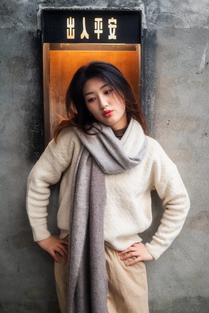 Young woman in a white sweater and gray scarf standing in front of a door.