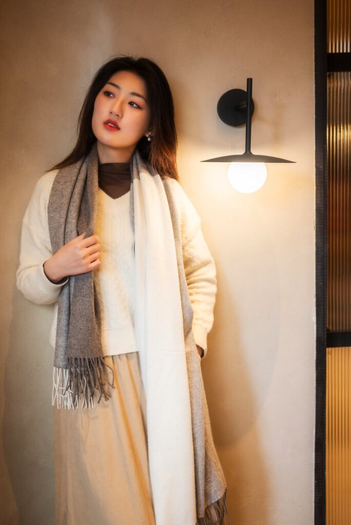 Young woman in a white sweater and gray scarf standing by the wall.