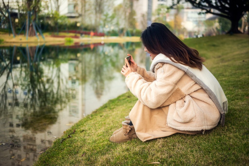 Young woman sitting on the grass by the lake using smartphone.