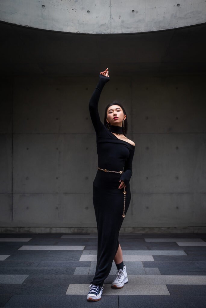 Young woman in black dress and sneakers posing in front of concrete wall