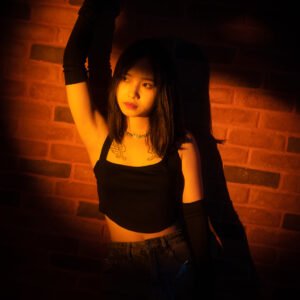 Young Asian woman standing in front of a brick wall with one arm raised.