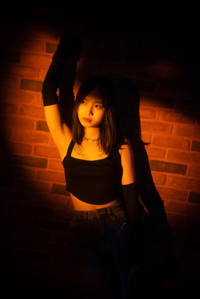 Young Asian woman standing in front of a brick wall with one arm raised.