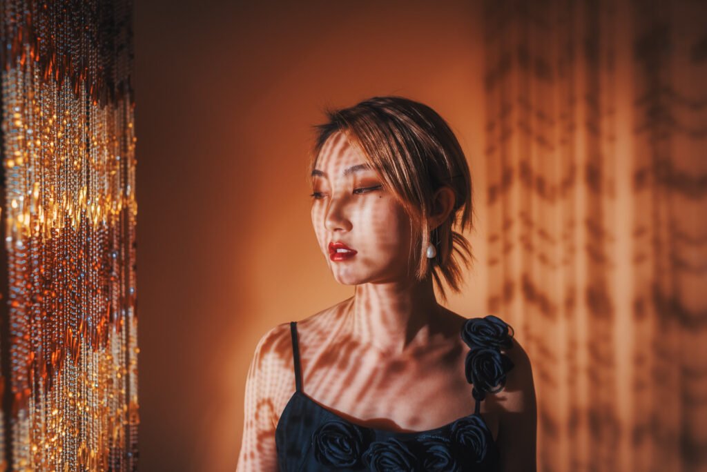 Portrait of a young woman standing in front of a curtain with sunlight and shadow on her face and body.